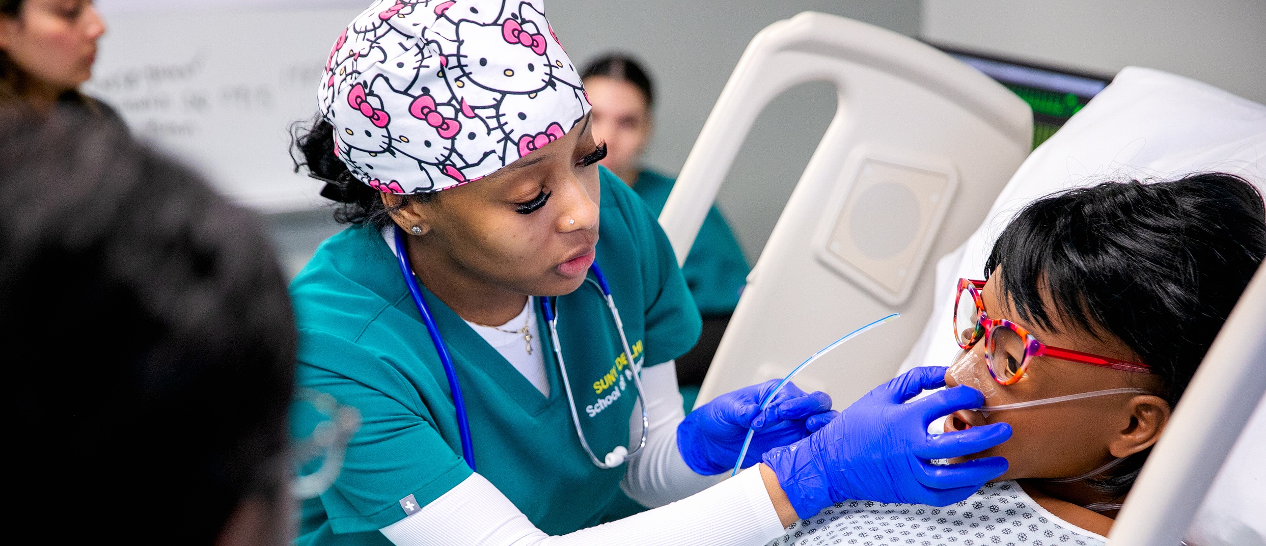 Nurse in teal scrubs helping a patient with oxygen tubes in a hospital bed.